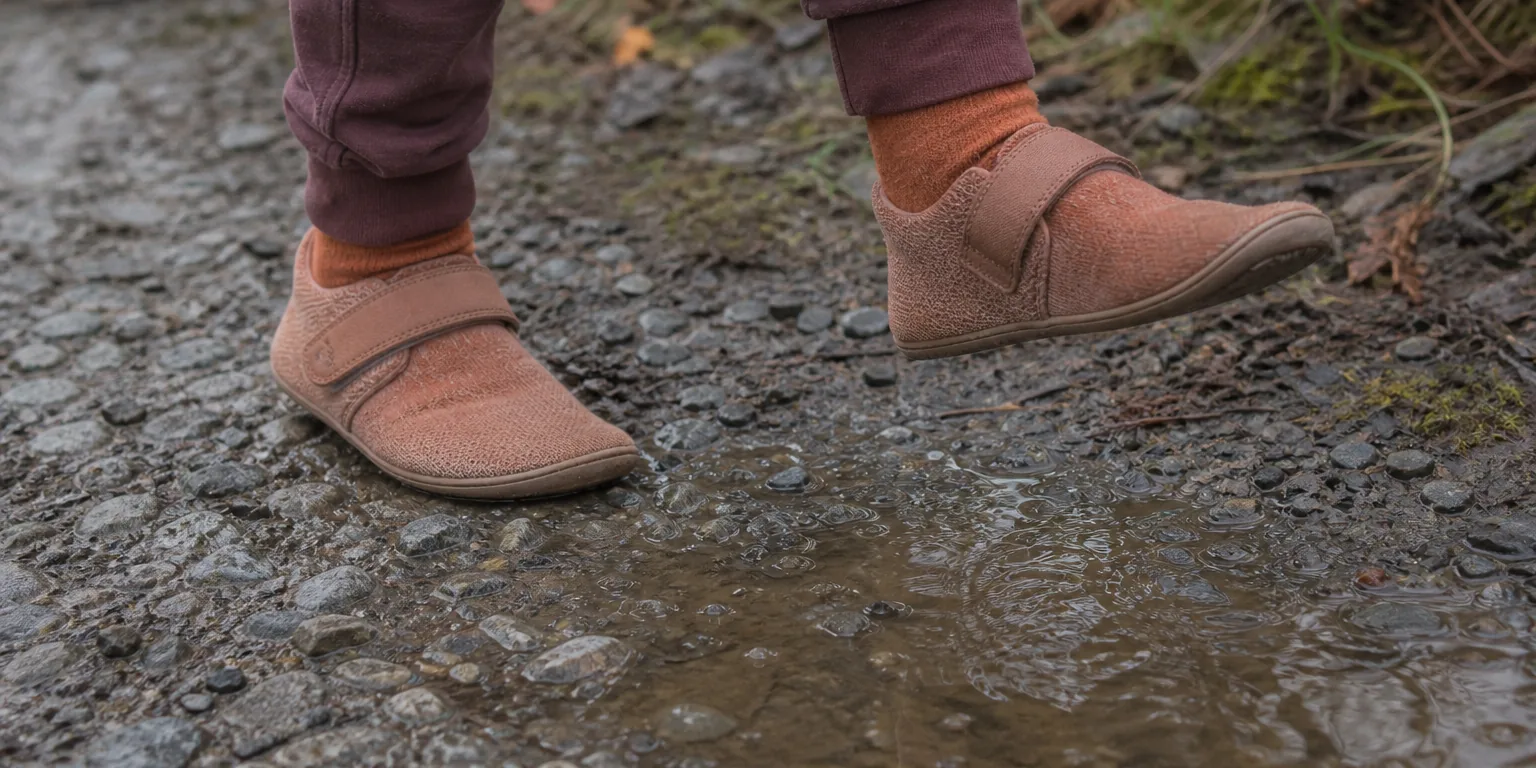 Child's feet in wide toe box barefoot shoes at the edge of a puddle on wet UK gravel