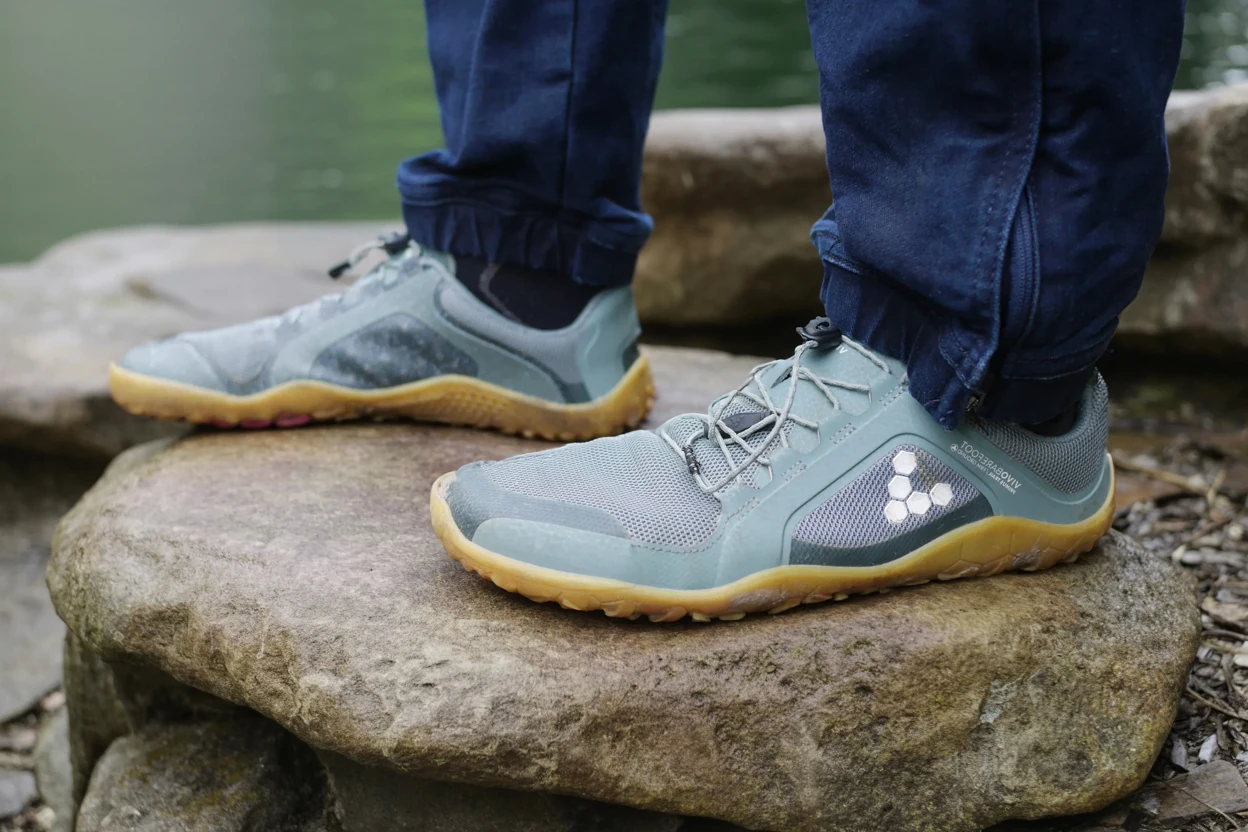 A person wearing a pair of barefoot shoes, vivobarefoot, a model that is recommended for plantar fasciitis. Barefoot minimalist running shoes on natural stone pathway showing wide toe box for plantar fasciitis recovery